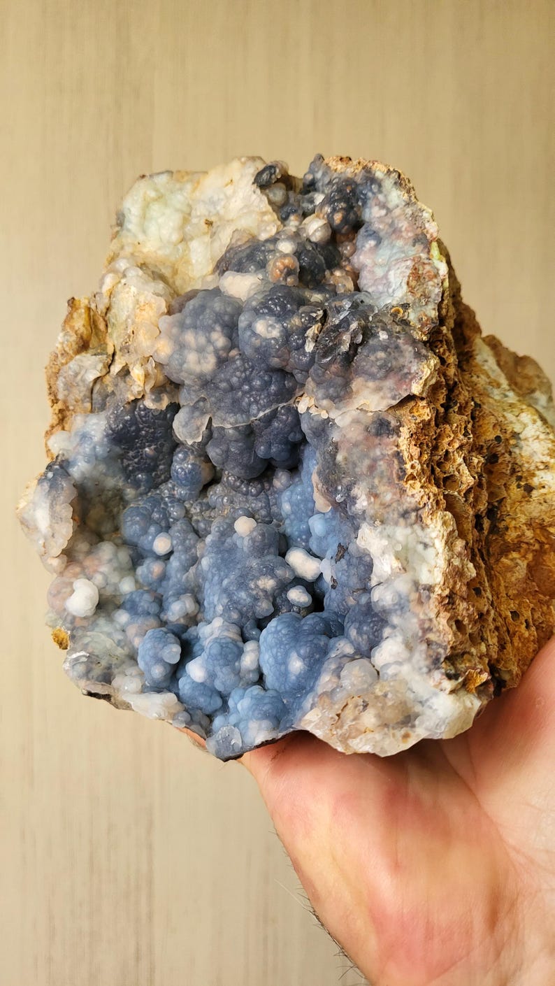 May include: A natural geode specimen with a cluster of blue chalcedony crystals. The geode has a rough, brown exterior and a cavity filled with small, rounded, blue-grey formations. The overall appearance is textured and organic.