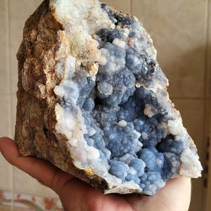 May include: A large geode with a rough, earthy exterior and a vibrant interior. The outer rock is a mix of brown and tan, contrasting with the light blue, spherical crystals within. The geode is held in a hand.
