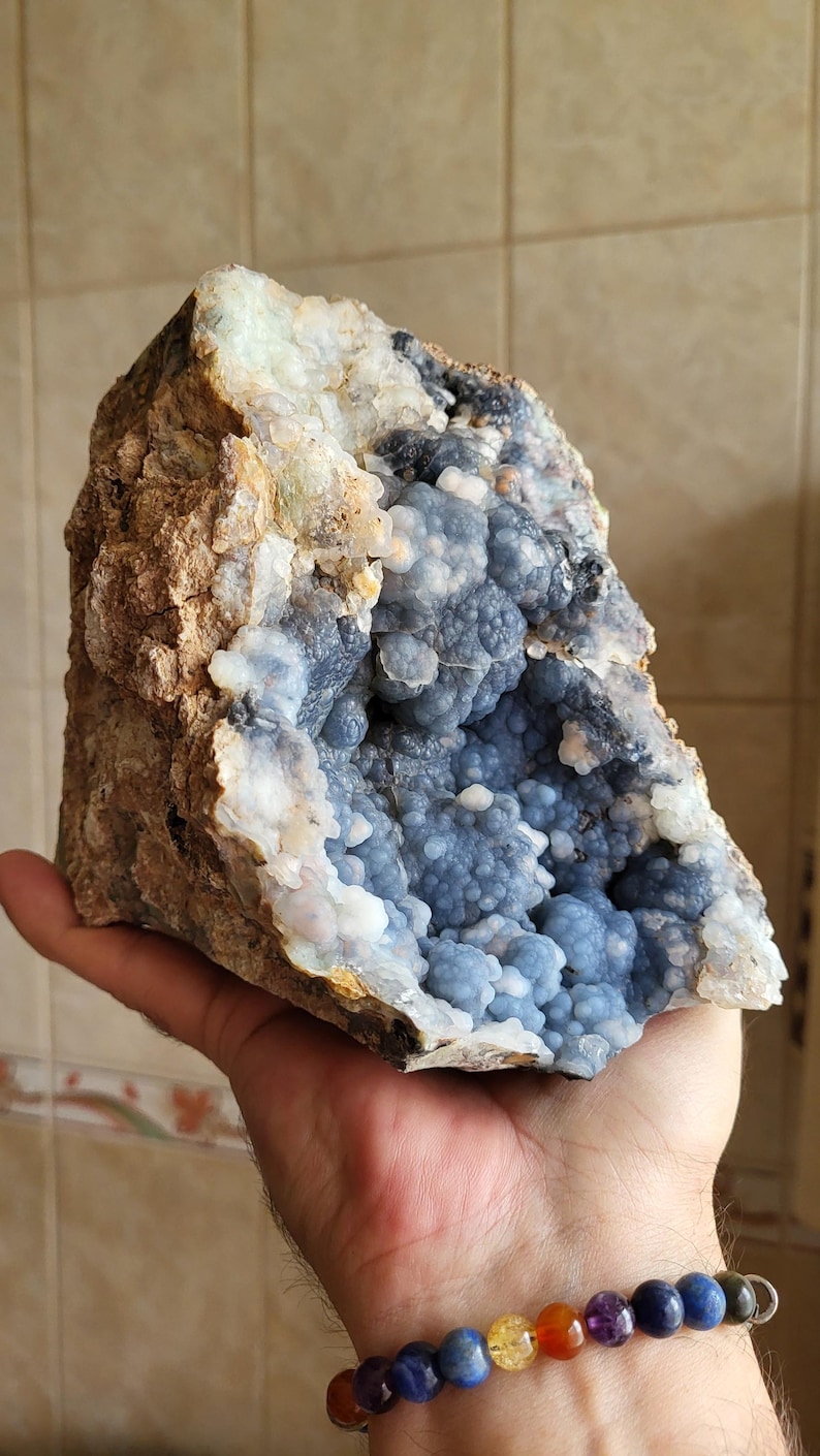 May include: A large geode with a rough, earthy exterior and a vibrant interior. The outer rock is a mix of brown and tan, contrasting with the light blue, spherical crystals within. The geode is held in a hand.