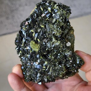 Peut inclure: Un spécimen minéral brut composé d'un amas dense de cristaux sombres, métalliques et aciculaires, entremêlés de formations cristallines vert olive éclatantes. La roche irrégulière présente une texture contrastée et brillante.