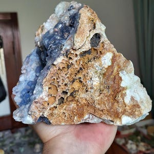 May include: A large, irregularly shaped rock formation with a mix of colours. The rock features areas of light blue, brown, and white, with a porous texture. The rock is held in a hand.