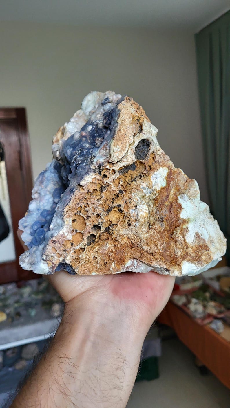 May include: A large, irregularly shaped rock formation with a mix of colours. The rock features areas of light blue, brown, and white, with a porous texture. The rock is held in a hand.