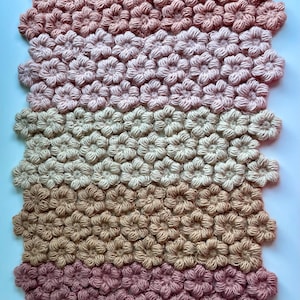 May include: A handmade, rectangular rug featuring a floral pattern. The rug is made of yarn in shades of pink, beige, and brown, creating a gradient effect. A small leather tag with text is attached to the bottom left corner.