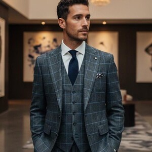 May include: A three-piece gray plaid suit with a white dress shirt and navy blue tie. The suit includes a blazer, vest, and trousers. A pocket square is visible in the blazer pocket. The suit is tailored and worn in a well-lit interior.