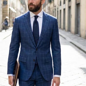 May include: A blue plaid suit with a white dress shirt and navy tie. The suit jacket has a two-button closure and the matching pants are tailored. A brown leather portfolio is held in the left hand. The man is walking down a cobblestone street.