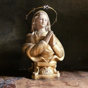 Puede incluir: Un busto de yeso pintado de oro de una mujer con un halo dorado y una corona de estrellas. La mujer lleva una larga túnica fluida y tiene las manos juntas en oración. El busto está sobre un pedestal con detalles ornamentados.