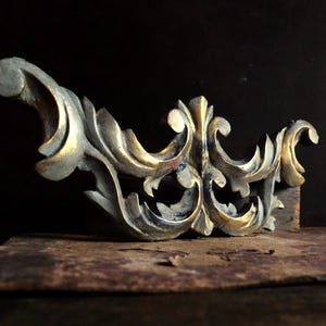 May include: A decorative wooden architectural element featuring intricate carved scrollwork and acanthus leaf motifs. The piece is finished with a distressed paint application, showing a combination of muted grey tones and worn gold leaf highlights.