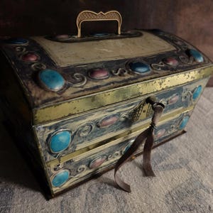 Puede incluir: Cofre de metal vintage con tapa abisagrada, adornado con piedras preciosas de imitación turquesas y rosas. Tiene un asa y un ribete dorados, con una cinta decorativa colgando de la cerradura. El exterior está pintado en tonos azules y crema.