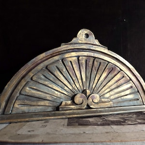 Puede incluir: Elemento arquitectónico decorativo semicircular con diseño de rayos de sol. La pieza es principalmente dorada con una apariencia desgastada y envejecida. Presenta detalles intrincados y volutas, lo que sugiere un estilo vintage o antiguo.