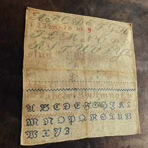 以下が含まれることがあります： ベージュのリネン生地に、アルファベット、数字1から9、そして「stud」という言葉が、青、緑、茶色の糸で刺繍されたサンプラーです。