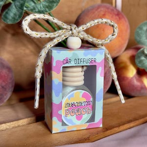 May include: A small car diffuser with a colourful design and the text "Enchanted Peach" on the front. The diffuser is packaged in a clear plastic box with a gold ribbon.