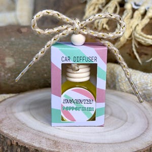 May include: A small, white box with a pink and green striped design. The box has a clear window showing a small glass bottle with a yellow liquid. The bottle is labelled "Scented Peppermint" and "Car Diffuser". The box is tied with a gold and white string.