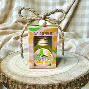 May include: A small, green and pink car diffuser with a wooden hanger. The diffuser is labelled "CAR DIFFUSER" and "ENCANTED VANILLA".