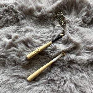 May include: Two miniature baseball bat keychains. One keychain has the word "NAME" engraved on it. The bats are light brown with black and white accents. The keychains are made of metal and are attached to a silver key ring.