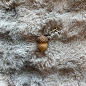 Può includere: Un piccolo portachiavi a forma di ghianda con la parte superiore marrone e la base marrone chiaro. Il portachiavi ha una chiusura e una catena color argento. Lo sfondo è un materiale morbido, grigio e peloso.