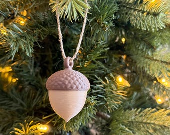 Può includere: Un ornamento natalizio a forma di ghianda, con una parte superiore marrone scuro e strutturata e una parte inferiore marrone chiaro. Appeso a uno spago naturale, si staglia su uno sfondo di un albero di Natale verde con luci bianche calde.