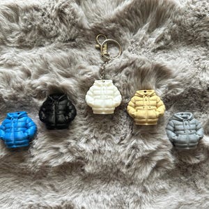 May include: A selection of miniature puffer jacket keyrings in blue, black, white, yellow, and grey. Each keyring features a detailed design of a hooded jacket with a zip and pockets. The keyrings are displayed on a soft, grey, furry surface.