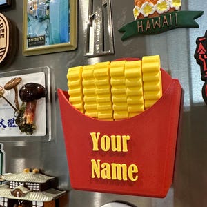 May include: A red and yellow 3D printed french fry holder magnet with the words "Your Name" in yellow letters. The magnet is attached to a stainless steel refrigerator.