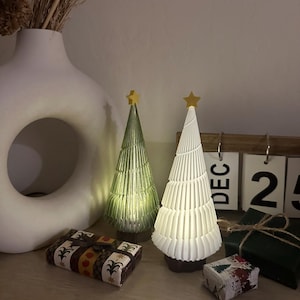Pode incluir: Duas árvores de Natal decorativas com uma estrela dourada no topo. A árvore à esquerda é verde e a árvore à direita é branca. Ambas as árvores são iluminadas com luzes. As árvores estão sentadas sobre uma superfície de madeira com presentes embrulhados.