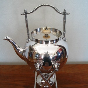 May include: Silver teapot with a bamboo-style handle and stand. The teapot has a round body and a lid with a knob. The stand has three legs that cross in the middle.
