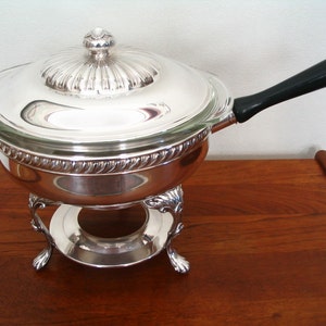 May include: Silver chafing dish with a glass lid and a black handle. The dish is on a stand with three legs.