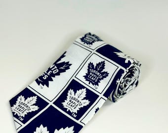 Pode incluir: Uma gravata com um padrão de grade repetitivo do logótipo dos Toronto Maple Leafs. O design consiste em quadrados alternados azul-escuro e brancos, cada um exibindo o emblema da folha de ácer branca e o texto Toronto Maple Leafs TM.