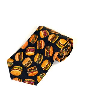 Peut inclure: Une cravate noire avec un motif répétitif de cheeseburgers. Chaque illustration de burger montre un pain aux graines de sésame, un steak haché, de la laitue verte, de la tomate rouge et des tranches d'oignon violet. La cravate est pliée en haut.