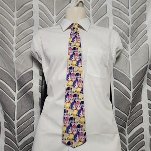 May include: A patterned necktie with a repeating cartoon character design in pink, purple, yellow, and blue. The tie is displayed on a white collared shirt.