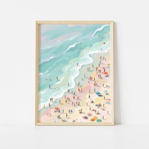 May include: A framed art print depicting a beach scene with numerous people. The artwork features a light blue ocean with white waves meeting a sandy shore. People are scattered across the beach, some with colorful umbrellas. The frame is a light wood color.