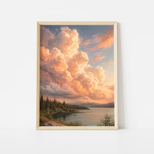 May include: A framed landscape painting depicting a serene lake at sunset. Large, voluminous clouds in shades of peach, orange, and soft blue dominate the sky above a dark green forest line and calm water, all encased in a light wood frame.