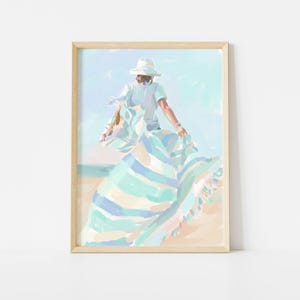 May include: A framed art print shows a person on a beach with a white hat and a striped blanket. The artwork uses pastel colours, including blue, green, and beige, creating a serene atmosphere. The print is in a light wood frame, suitable for a coastal theme.
