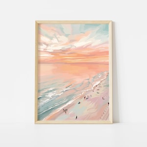 May include: A framed art print featuring a stylised beach scene at sunset with soft pastel hues of peach, pink, and light blue. The composition shows a sandy shoreline with small figures of people scattered along the water's edge under a cloudy sky.