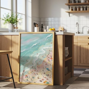 May include: A framed art print depicts a beach scene with turquoise water, a sandy shore, and numerous tiny figures of people. The artwork has a light wooden frame and is displayed on a kitchen island. The style is reminiscent of a painting.