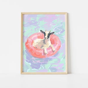 May include: A framed art print depicts a white goat with black markings, relaxing on a pink inflatable ring in water. The water has a colorful, abstract design in shades of blue, green, and purple. The artwork is framed in a light wood.