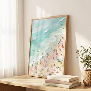 May include: A framed art print depicts a vibrant beach scene. Turquoise waves meet a sandy shore dotted with numerous figures, umbrellas, and beach towels. The artwork is encased in a light-colored wooden frame, resting on a wooden surface with books and a plant.