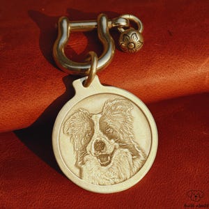 May include: A gold-coloured dog tag featuring an engraved portrait of a dog. The tag is connected to a gold-coloured clasp and a small decorative charm. The background is a reddish-brown leather surface.
