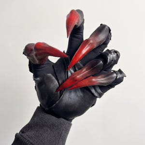 May include: A black glove featuring long, pointed, claw-like extensions on each finger. The claws transition from a deep black base to a vibrant, blood-red tip. The glove is constructed from a smooth, matte black material with a ribbed cuff at the wrist.