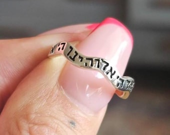 May include: A silver-toned metal ring featuring a wavy, undulating band design. The surface of the ring is engraved with black Hebrew text that follows the curved contour of the metal, creating a distinct visual pattern along the entire band.