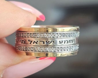 May include: A gold and silver ring with Hebrew text and small clear stones. The ring has multiple bands, with the text on one band. The ring is held by a person with pink nail polish.