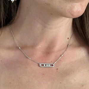 May include: A silver necklace with a rectangular pendant holding three small, clear stones. A gold earring with a green stone and three dangling pearls is visible in the upper left corner. The jewelry is worn on a person.