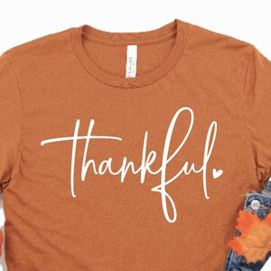 Peut inclure: Un t-shirt couleur rouille avec le mot "thankful" &eacute;crit en blanc en cursive. Le t-shirt est associ&eacute; &agrave; un jean bleu, des baskets blanches, une tasse blanche avec le texte "pumpkin spice" et des d&eacute;corations d'automne.
