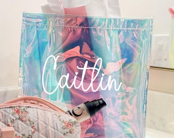 May include: A clear iridescent tote bag with a white handle and the name "Caitlin" printed on the front. The bag is filled with a pink and white striped towel. A small quilted floral makeup bag with a pink zipper is next to the tote bag. A clear bottle of lotion is inside the tote bag.