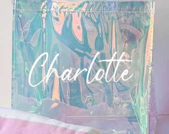 May include: A clear iridescent tote bag with the name "Charlotte" printed on it. A pink and white striped beach towel is rolled up next to the bag.
