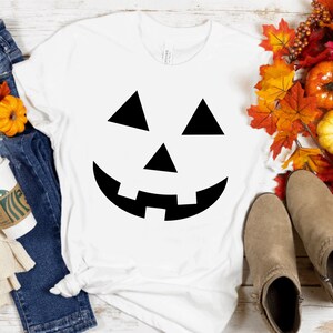 May include: A white short-sleeved t-shirt featuring a black jack-o'-lantern face graphic, paired with blue denim jeans, a beige scarf, a white coffee cup, tan ankle boots, and a decorative arrangement of colorful autumn leaves and pumpkins.