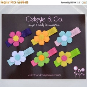 May include: A collection of colorful flower hair clips on a dark background. Each clip features a felt flower in various colors like pink, orange, blue, and green, attached to a matching ribbon. The brand name "Celeste & Co." is visible.
