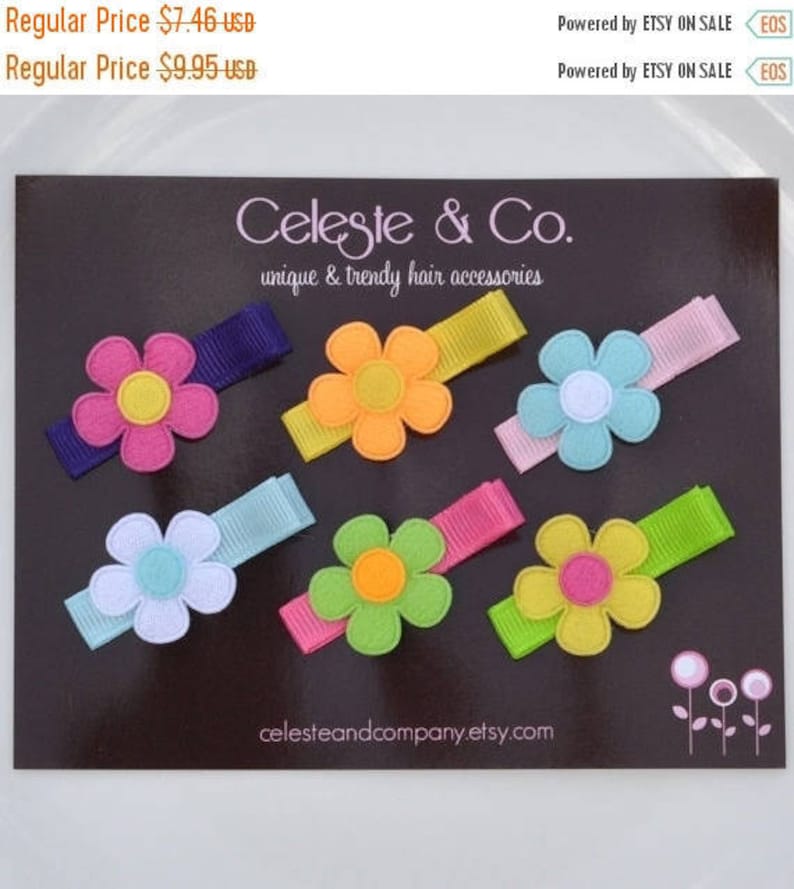 May include: A collection of colorful flower hair clips on a dark background. Each clip features a felt flower in various colors like pink, orange, blue, and green, attached to a matching ribbon. The brand name "Celeste & Co." is visible.
