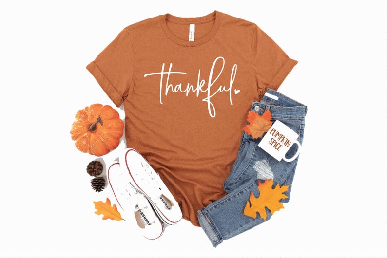 Peut inclure: Un t-shirt couleur rouille avec le mot "thankful" &eacute;crit en blanc en cursive avec un petit c&oelig;ur. Le t-shirt est pos&eacute; sur un fond blanc avec une paire de baskets blanches, une petite citrouille orange et une tasse blanche avec les mots "pumpkin spice" &eacute;crits en orange.
