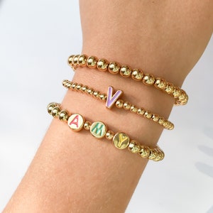 Puede incluir: Tres pulseras de cuentas doradas con abalorios de letras de colores que deletrean el nombre "AMY" y una sola pulsera de cuentas doradas con un abalorio de letra p&uacute;rpura "V".