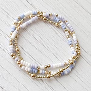 Puede incluir: Tres pulseras de cuentas apiladas. Las pulseras presentan una combinación de cuentas doradas, cuentas blancas y cuentas azul claro. Las pulseras están dispuestas sobre una superficie de madera de color claro.
