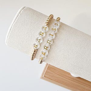May include: Two beaded bracelets with clear beads and gold lettering spelling "NANA" and "ABRISOS". The bracelets have gold accent beads. The bracelets are displayed on a white, cylindrical surface.